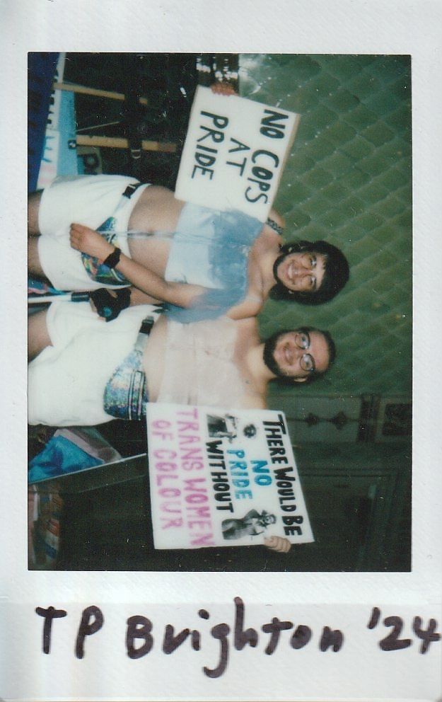Two individuals hold protest signs advocating for Pride inclusion and celebrating trans women of color.