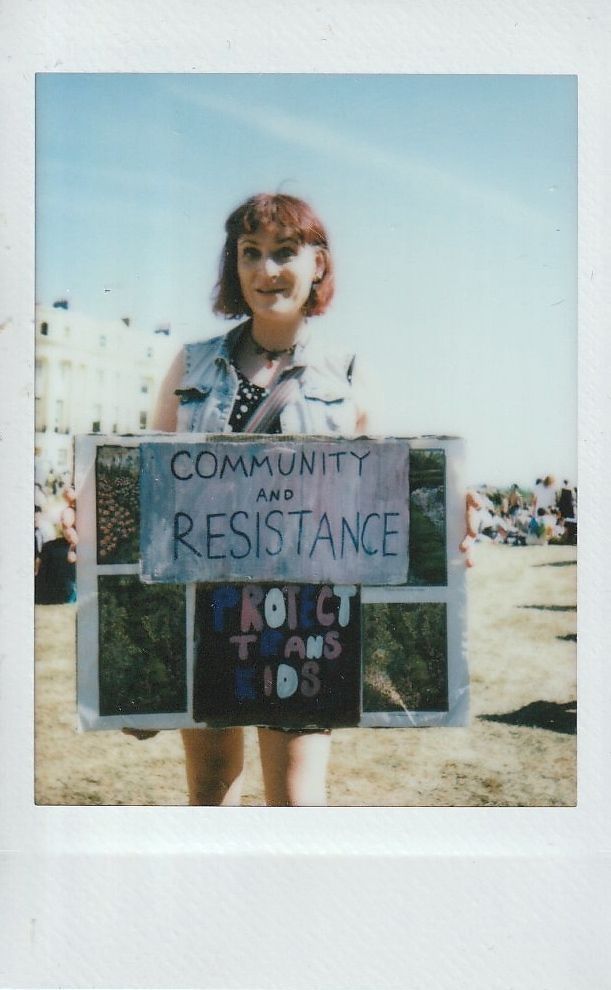 A person stands holding a sign which says "COMMUNITY AND RESISTANCE PROTECT TRANS KIDS".