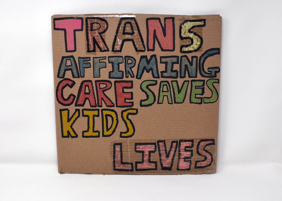 A placard made from cardboard which has 'Trans Affirming Care Saves Lives' in colourful block text.