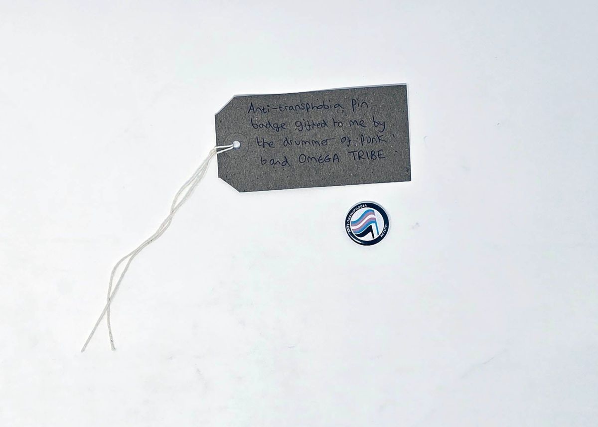 A badge with a black border and a trans flag within it and a brown luggage tag with writing on it.