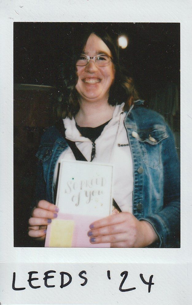 A person smiling holds a card reading "So Proud of You," wearing a denim jacket in Leeds '