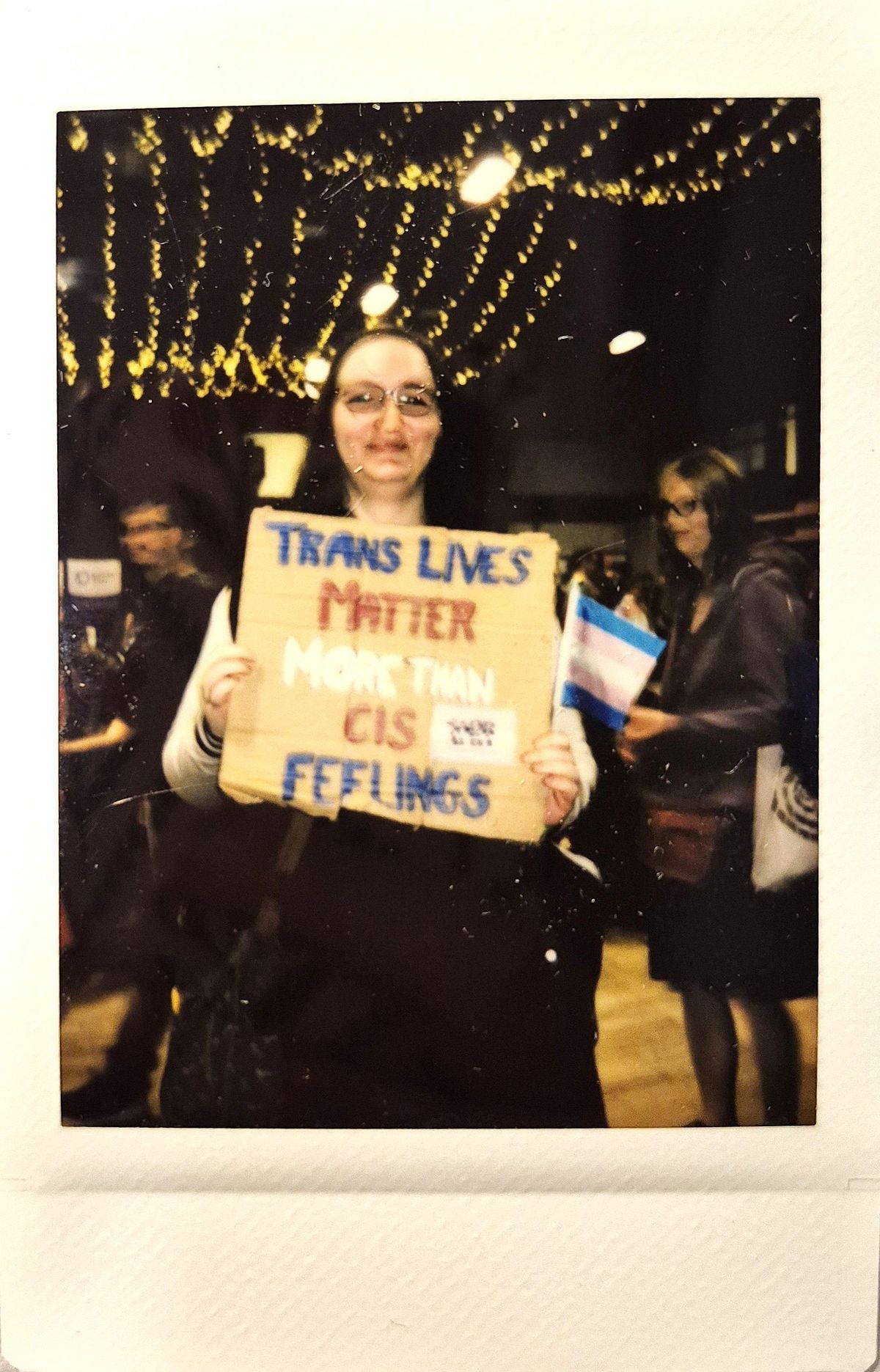 A person holds a sign saying "Trans Lives Matter More Than Cis Feelings" at an indoor gathering.