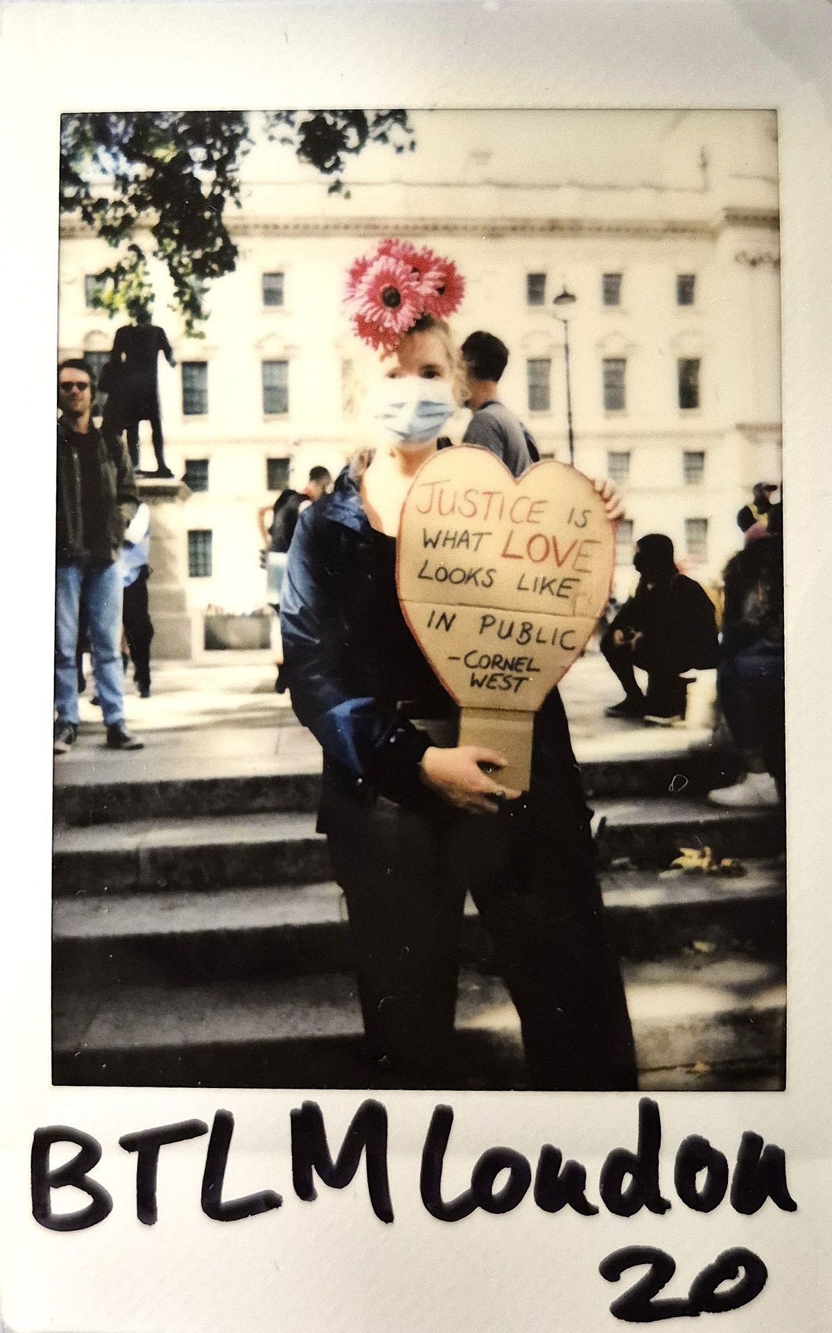 A person wearing a mask and flower headdress holds a heart-shaped sign.