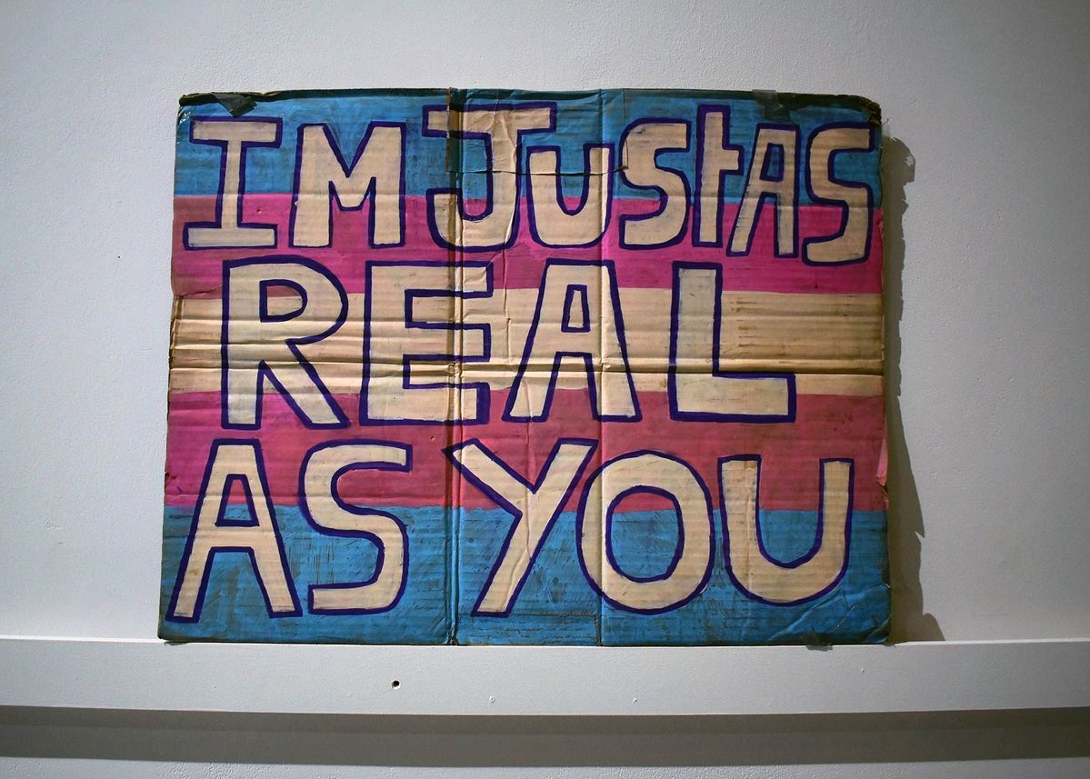 A hand painted cardboard placard in the trans flag colours and the text "IM JUST AS REAL AS YOU"