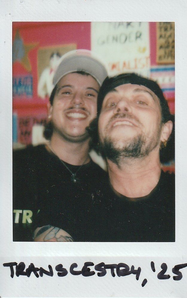 Two people are smiling at the camera in a colorful room with various posters in the background.