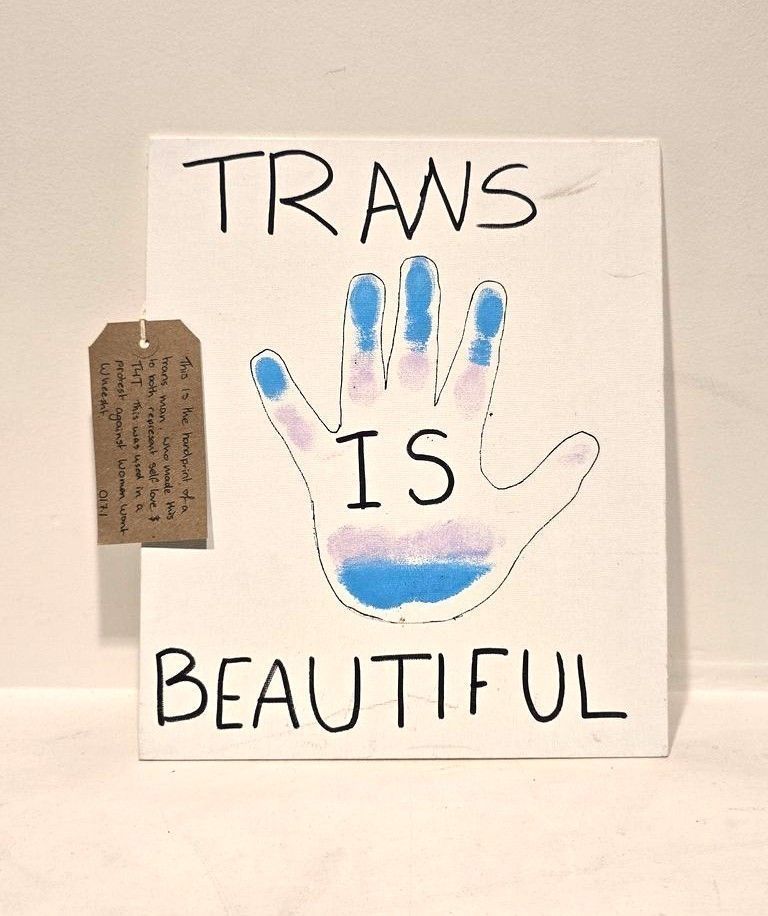 A placard which is white, has an outline of a hand drawn and inside is painted in trans flag colours - blue, pink and white. It has 'Trans is Beautiful' written on it. Lay on the placard is a brown tag with writing on it.
