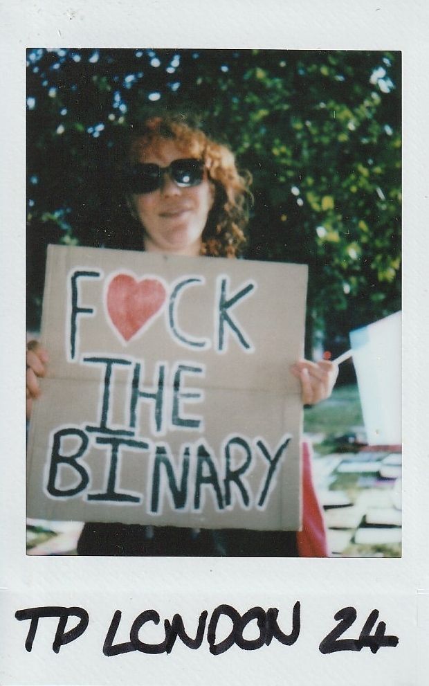 A person wearing sunglasses is holding a sign reading "F❤️CK THE BINARY".