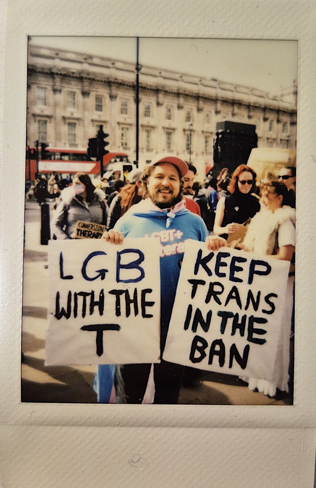 A person at a protest holds signs which say "LGB WITH THE T" and "KEEP TRANS IN THE BAN".