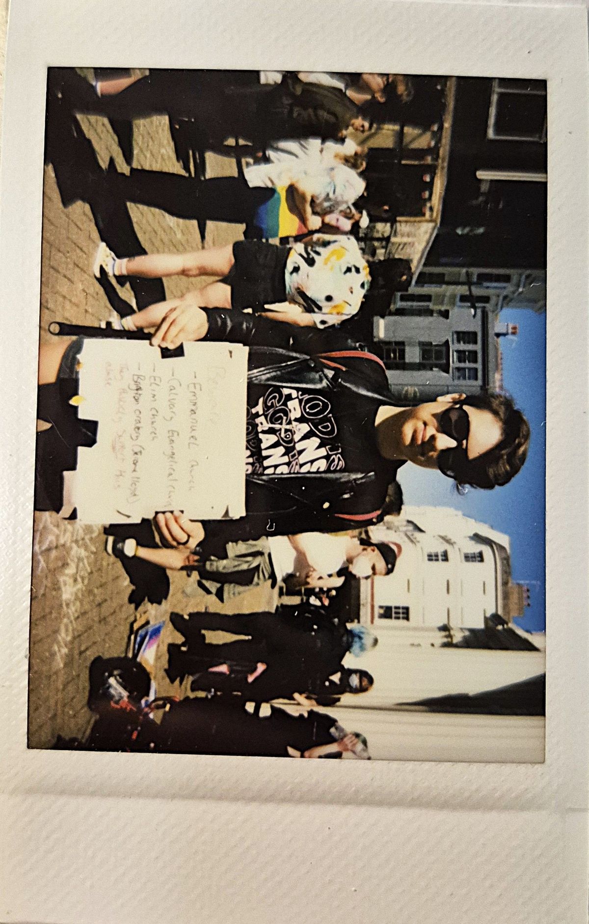 A person in sunglasses holds a handwritten sign during a sunny outdoor gathering with several people.