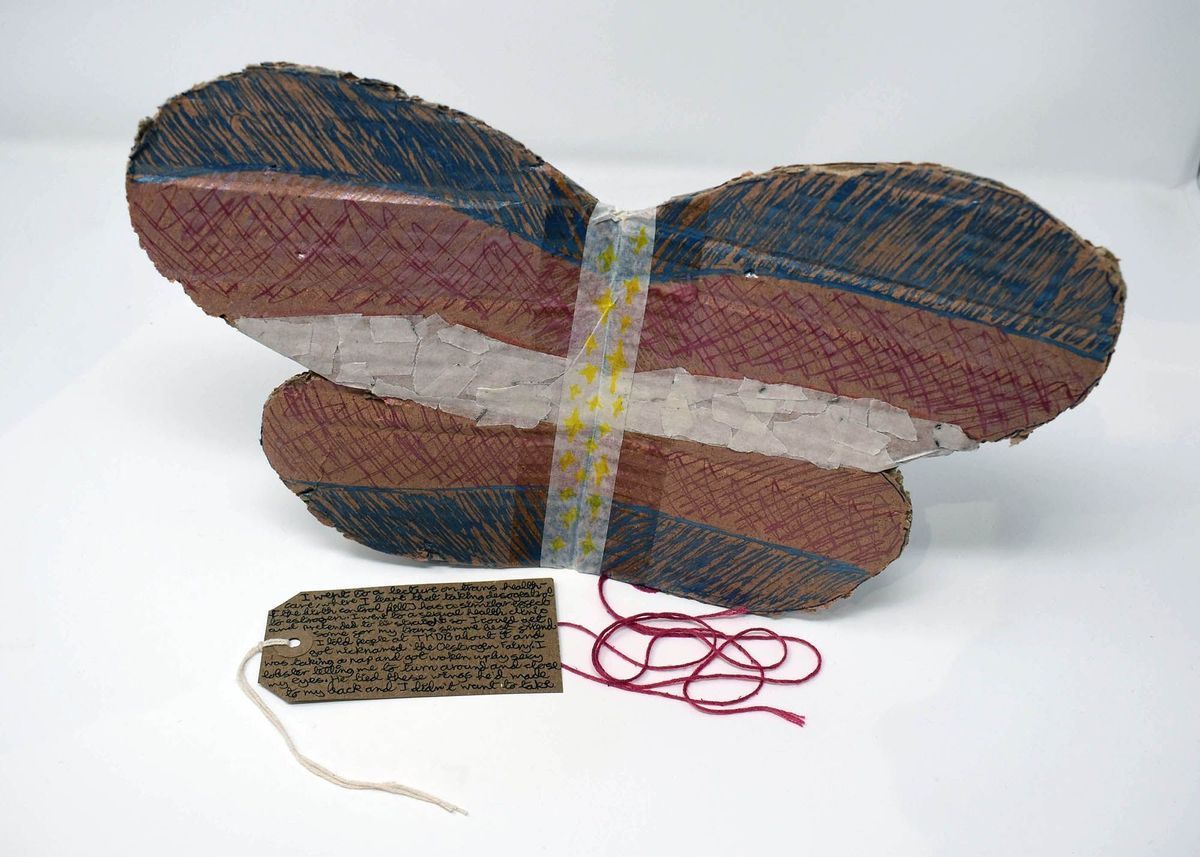 A pair of handmade cardboard fairy wings. They are coloured in blue, white and pink. With some red string and a brown, handwritten luggage tag.
