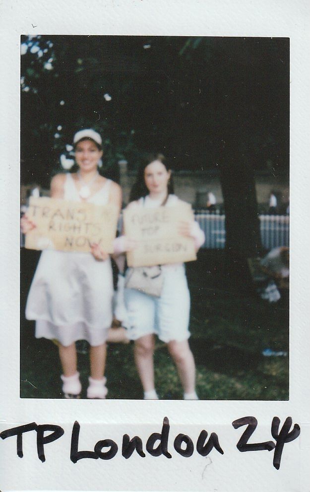 Two people holding protest signs stand together outdoors, one sign says "Trans Rights Now" and the other is too blurry to make out.