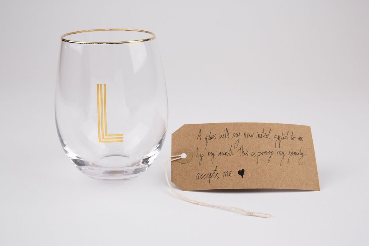 A small drinking glass with a gold rim and the letter "L" in gold. With a brown, handwritten luggage tag.