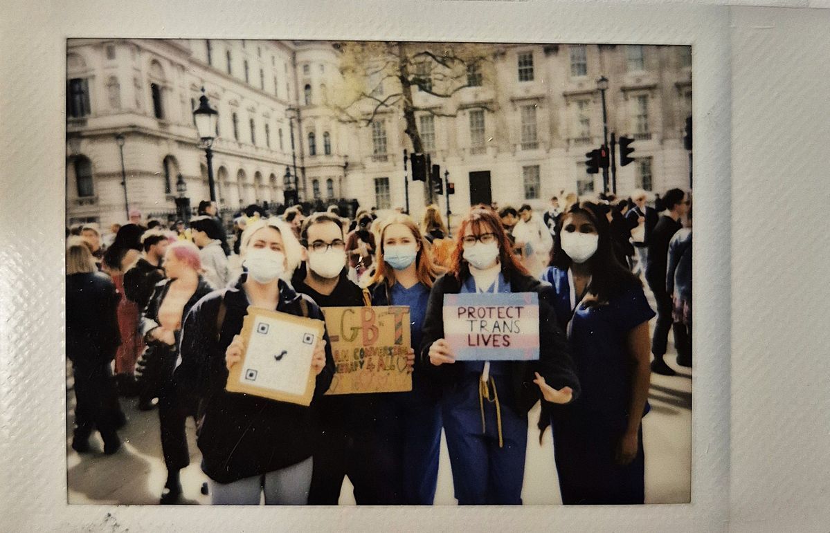 A group of people, wearing masks, hold signs saying phrases like "Protect Trans Lives".