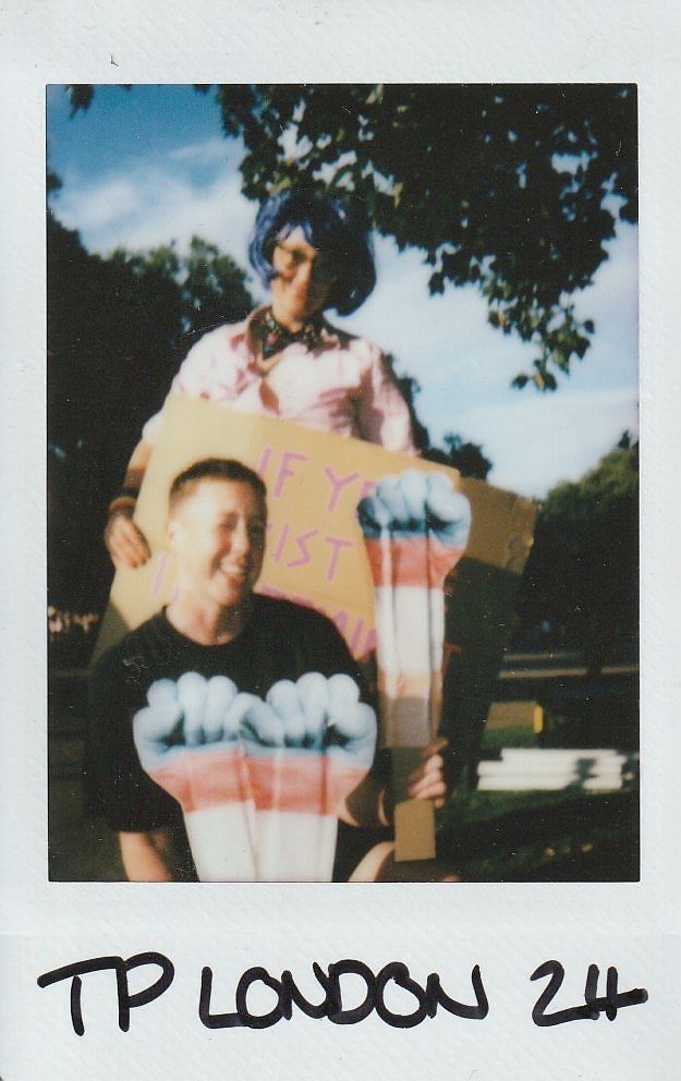 In the image, two people are outdoors holding signs with creative artwork, surrounded by trees.