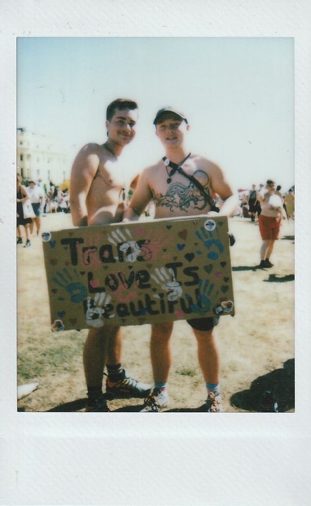 Two people smiling, holding a colorful sign that reads "Trans Love Is Beautiful".