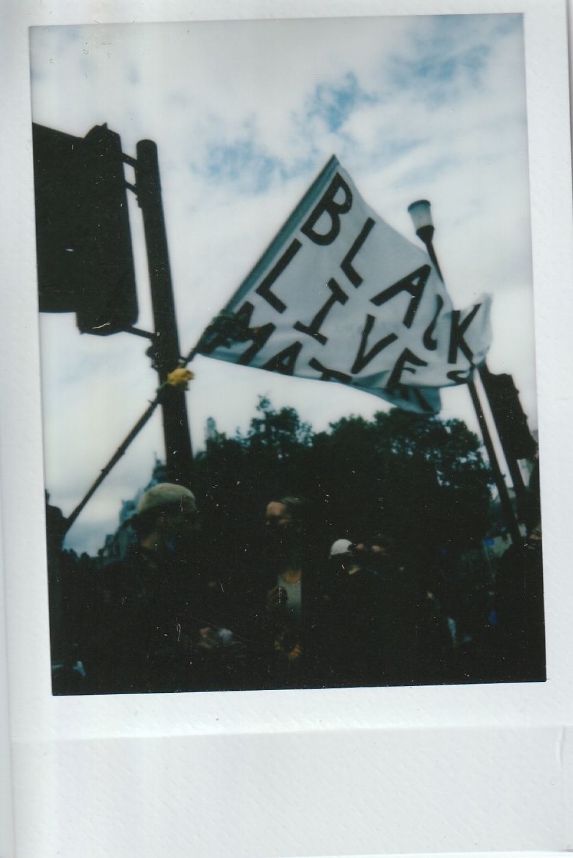 A group of people gathers under a "Black Lives Matter" flag, demonstrating in daylight.