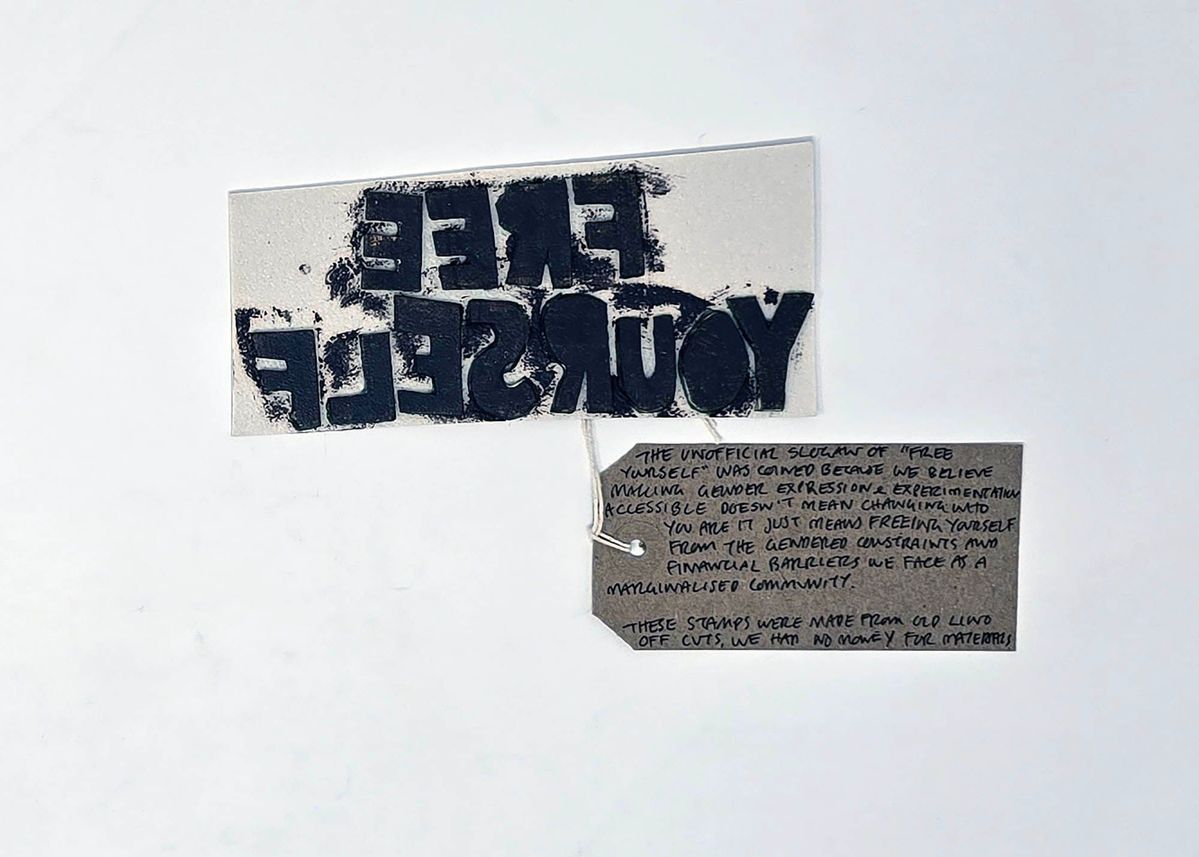 The image shows a reversed "FREE YOURSELF" lino cut stamp covered in black ink. With a brown, handwritten luggage tag.