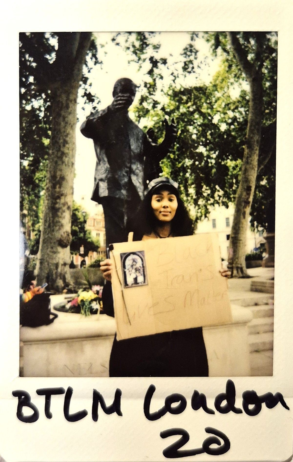 A person holds a sign reading "Black Trans Lives Matter" in front of a statue with trees behind.