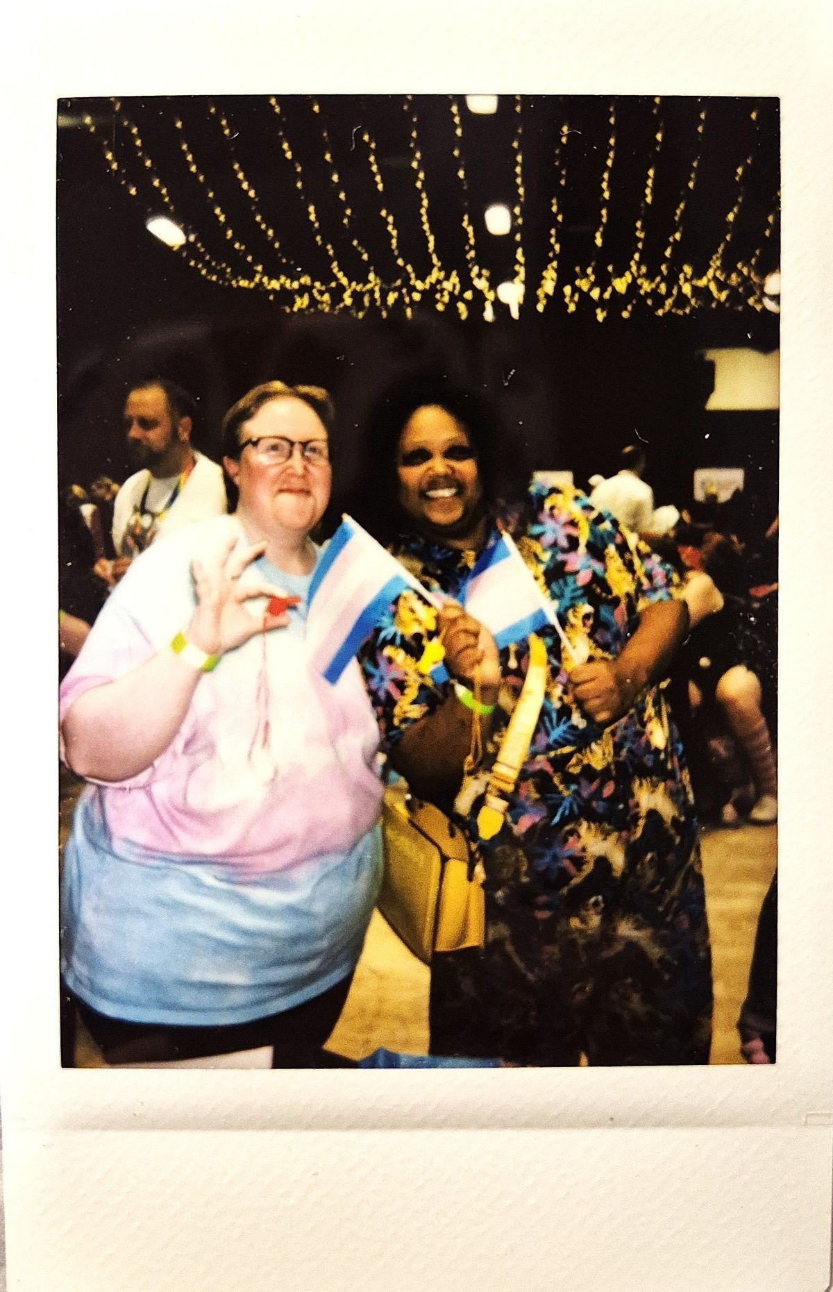 Two smiling individuals hold small transgender flags under decorative lights at a lively indoor event.