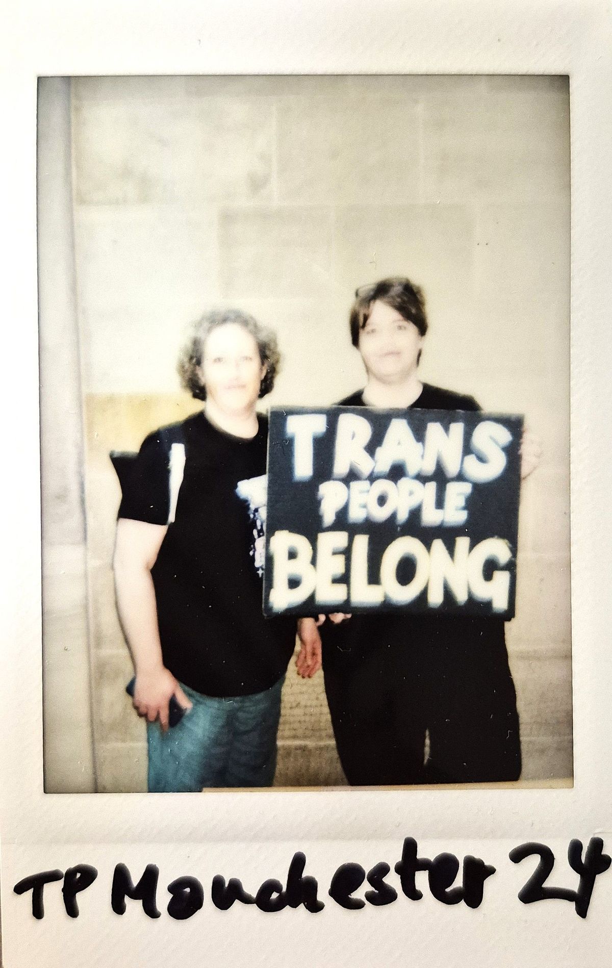 Two individuals, in black attire, hold a sign that says “Trans People Belong”.
