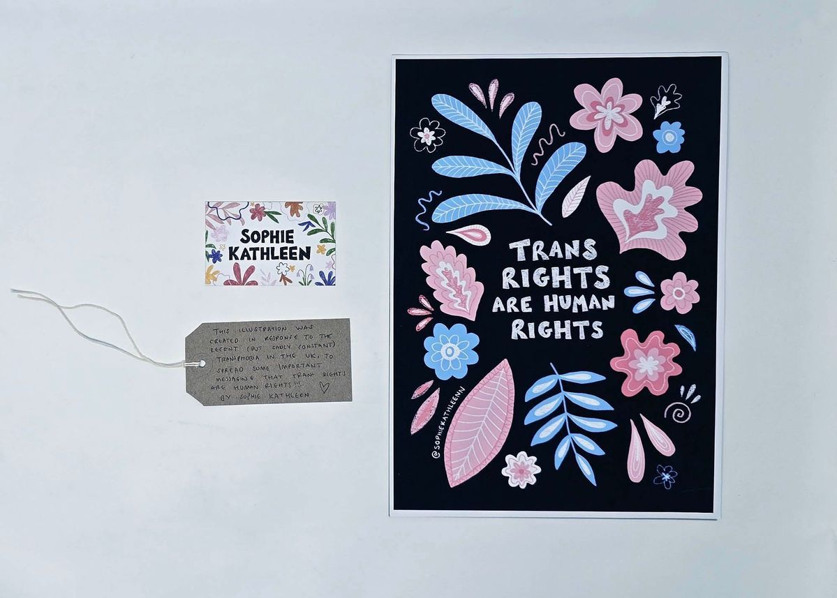 A print showing the text "TRANS RIGHTS ARE HUMAN RIGHTS" in white on a black background decorated with pink, white and blue leaves and flowers. With a name card, SOPHIE KATHLEEN, decorated with flowers, and a brown, handwritten luggage tag.