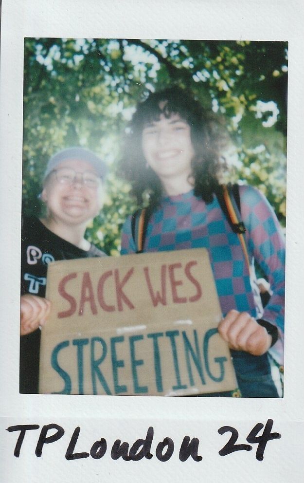 Two people are smiling and holding a "Sack Wes Streeting" sign, standing under leafy trees.