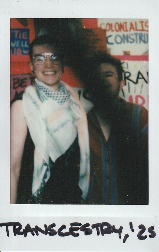 Two people are smiling in front of colorful signs, with "Transcestry, '23".