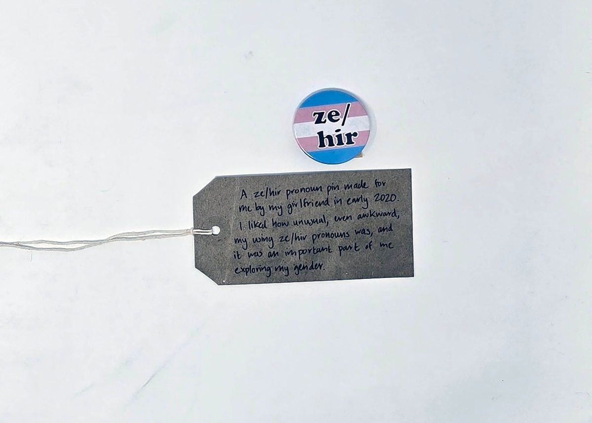 A small, round badge with black text"ze/hir" over a background in the trans pride colours. With a brown, handwritten luggage tag.