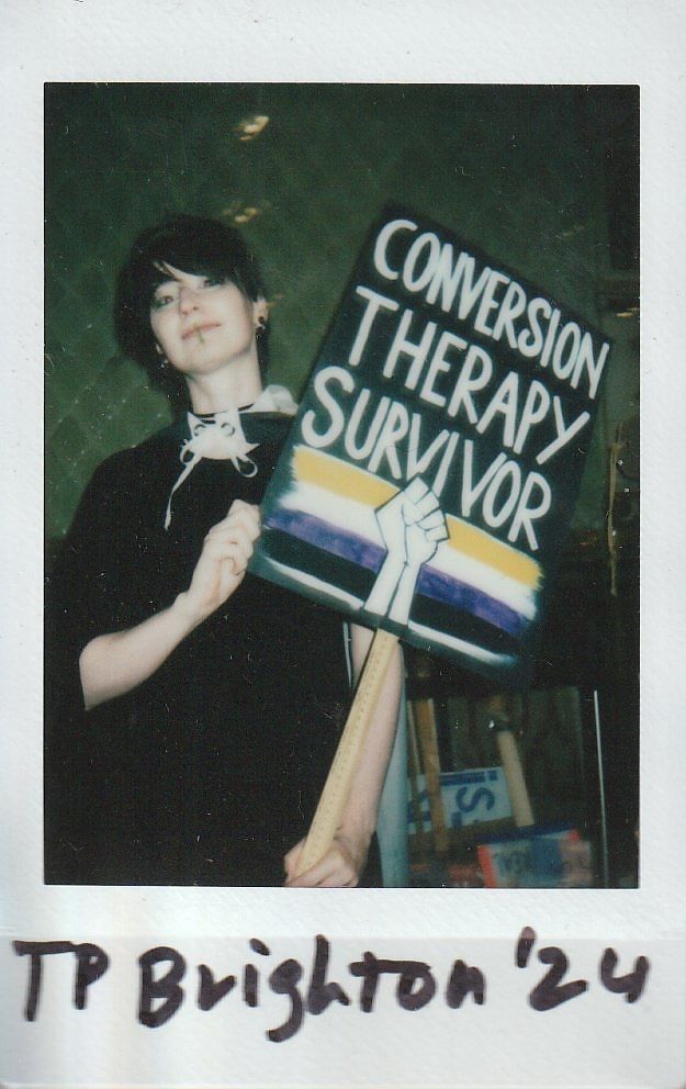 A person holds a sign reading "Conversion Therapy Survivor" with a rainbow flag design.