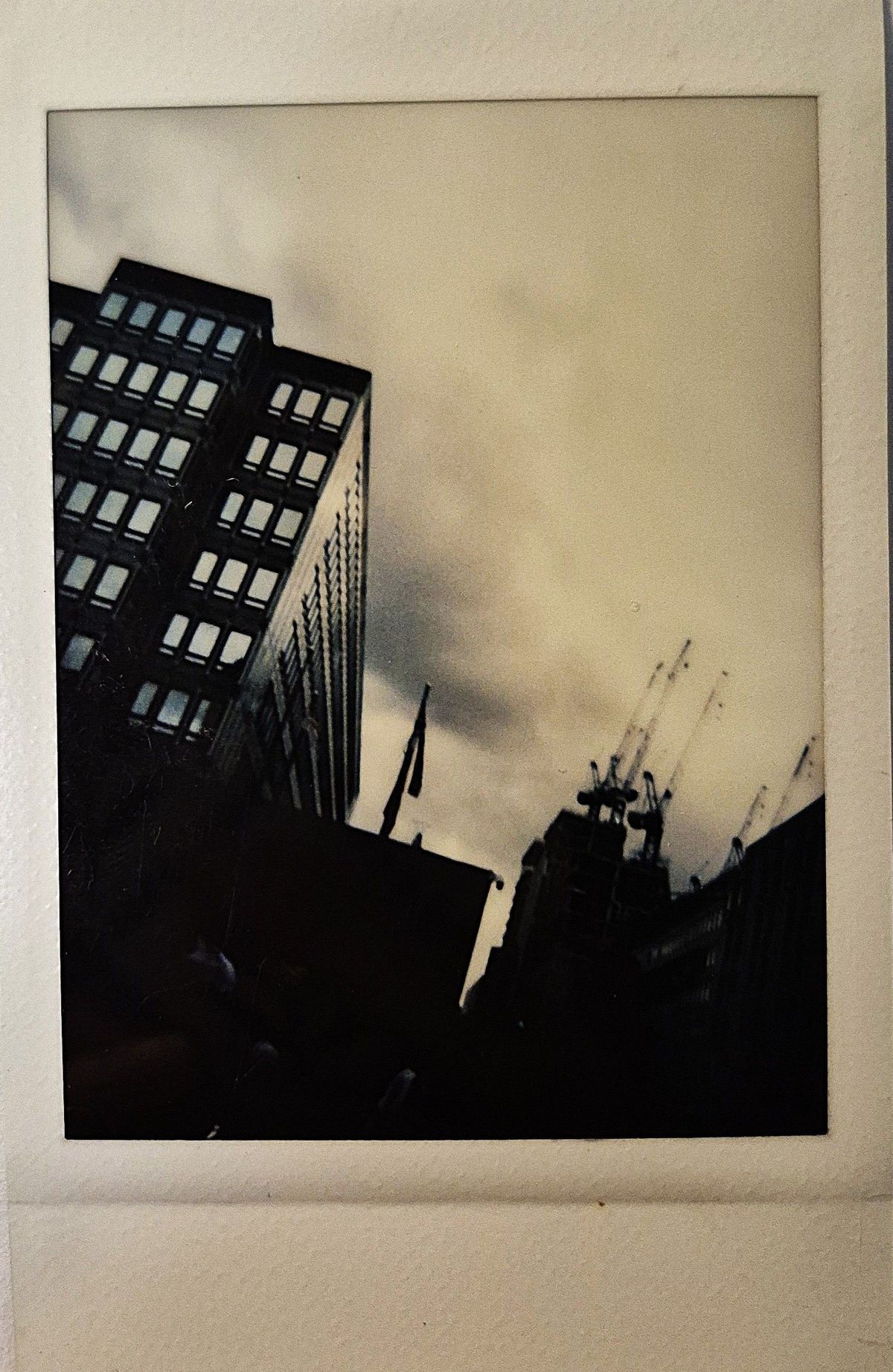 The image shows tall buildings and cranes silhouetted against an overcast sky, captured in a vintage photo