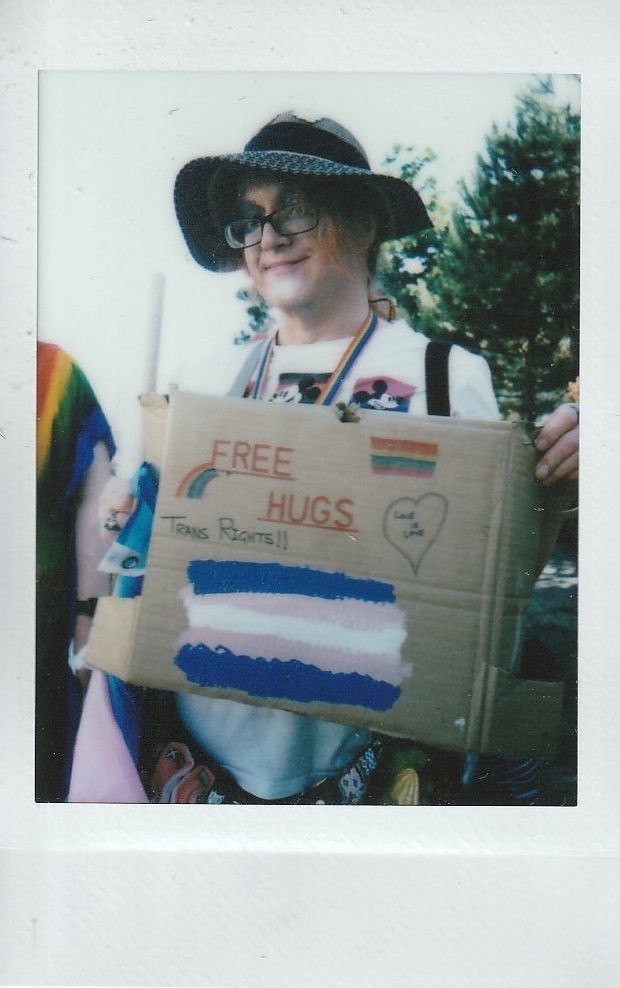 A person holds a "Free Hugs" wearing a hat and rainbow accessories.