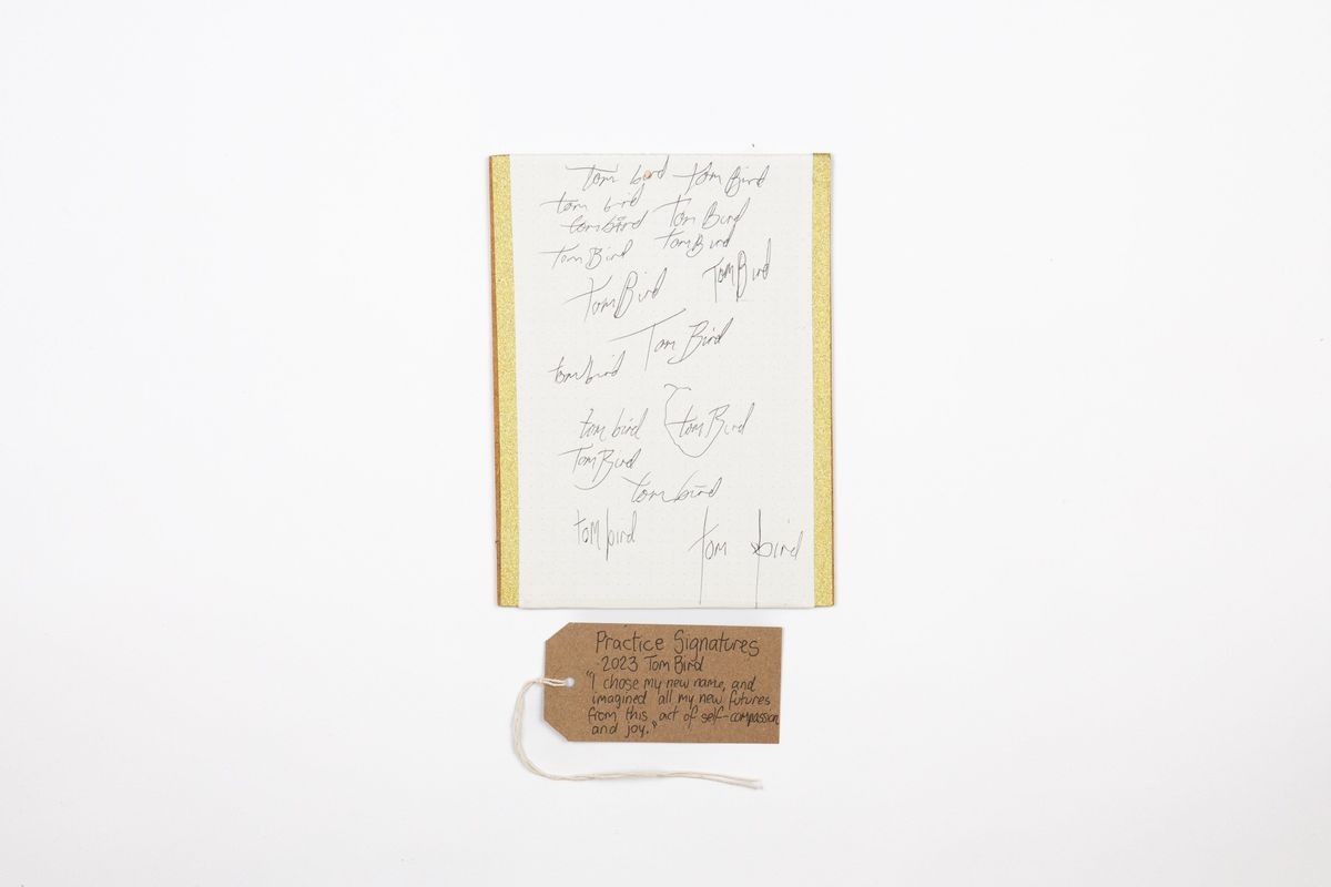 The image shows a sheet with handwritten practice signatures and a brown tag with writing on it sat underneath.
