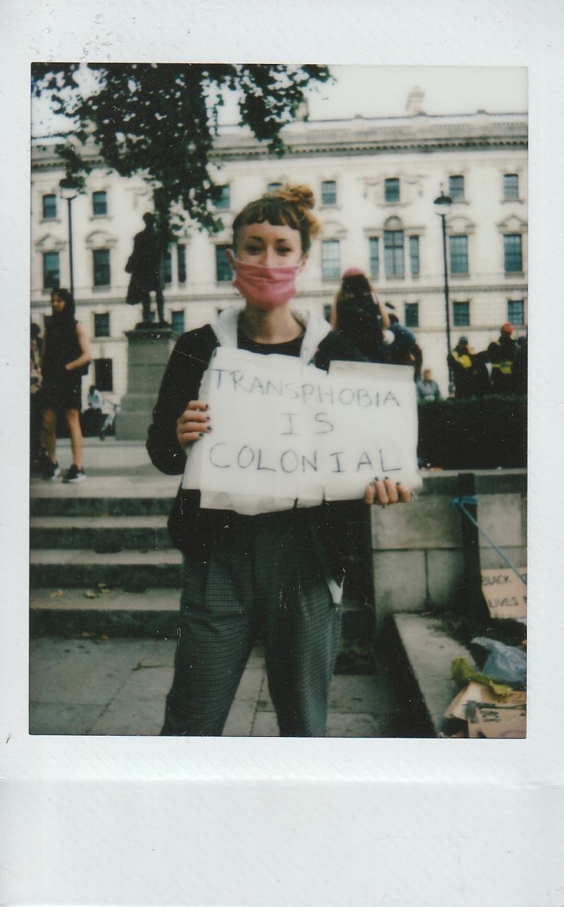 A person in a pink mask holds a sign that says, "Transphobia is Colonial".