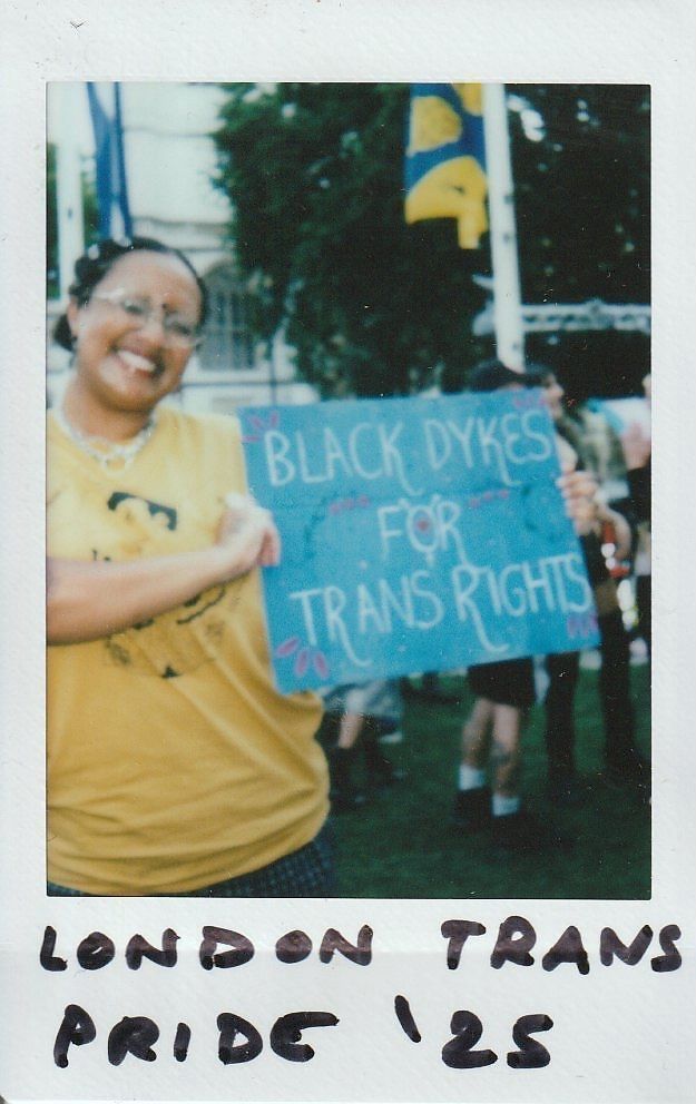 A person in a yellow shirt holds a sign which says "Black Dykes For Trans Rights".