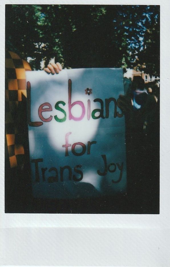A sign reading "Lesbians for Trans Joy" is held outdoors with trees visible.