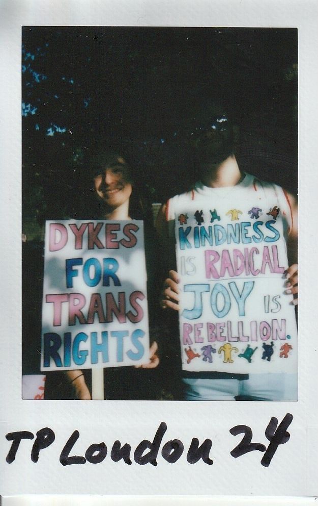 Two people hold signs saying "Dykes for Trans Rights" and "Kindness is Radical, Joy is Rebellion".