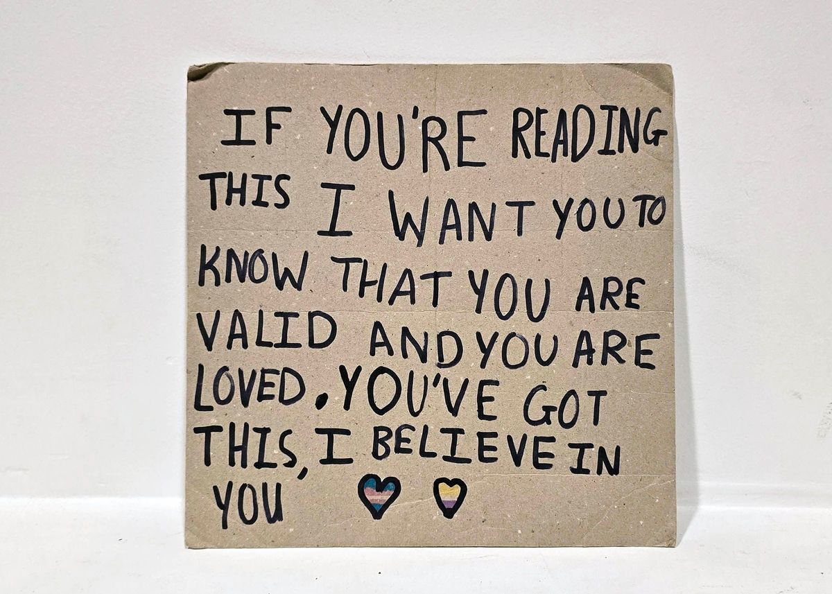 Handwritten cardboard placard with text "IF YOU'RE READING THIS I WANT YOU TO KNOW THAT YOU ARE VALID AN YOU ARE LOVED. YOU'VE GOT THIS, I BELIEVE IN YOU" followed by two hearts coloured in the trans and non-binary colours.