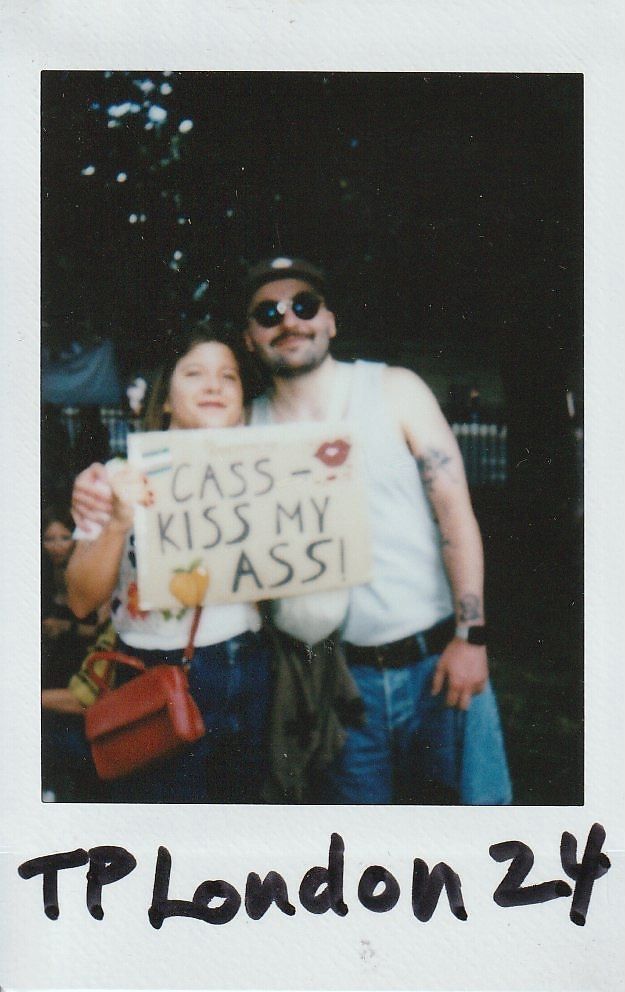 Two people stand together outdoors, holding a protest sign which says "CASS Can Kiss My Ass".