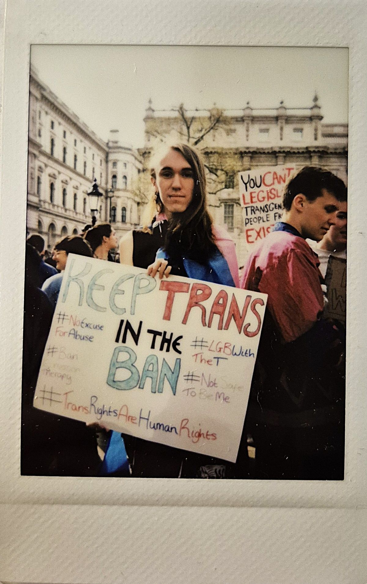 A person holds a sign which says "KEEP TRANS IN THE BAN" and various hashtags such as #TransRightsAreHumanRights.