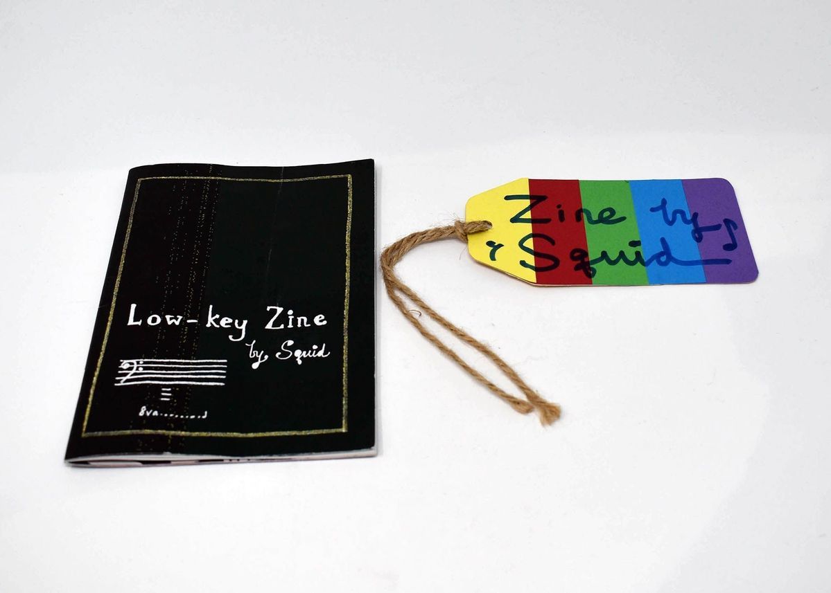 A black zine which says 'Low Key Zine by Squid' on the front. Next to the zine is a tag which is rainbow coloured and says in black marker 'Zine Squid'.