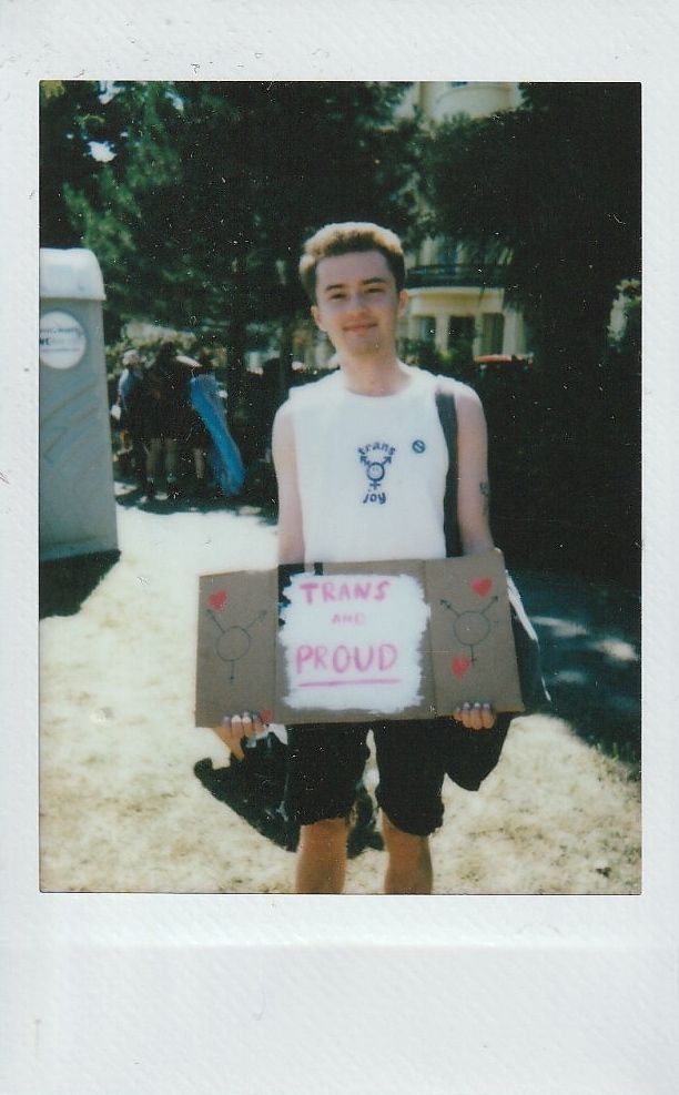 In a sunny outdoor setting, a person holds a "Trans and Proud" sign, smiling confidently.