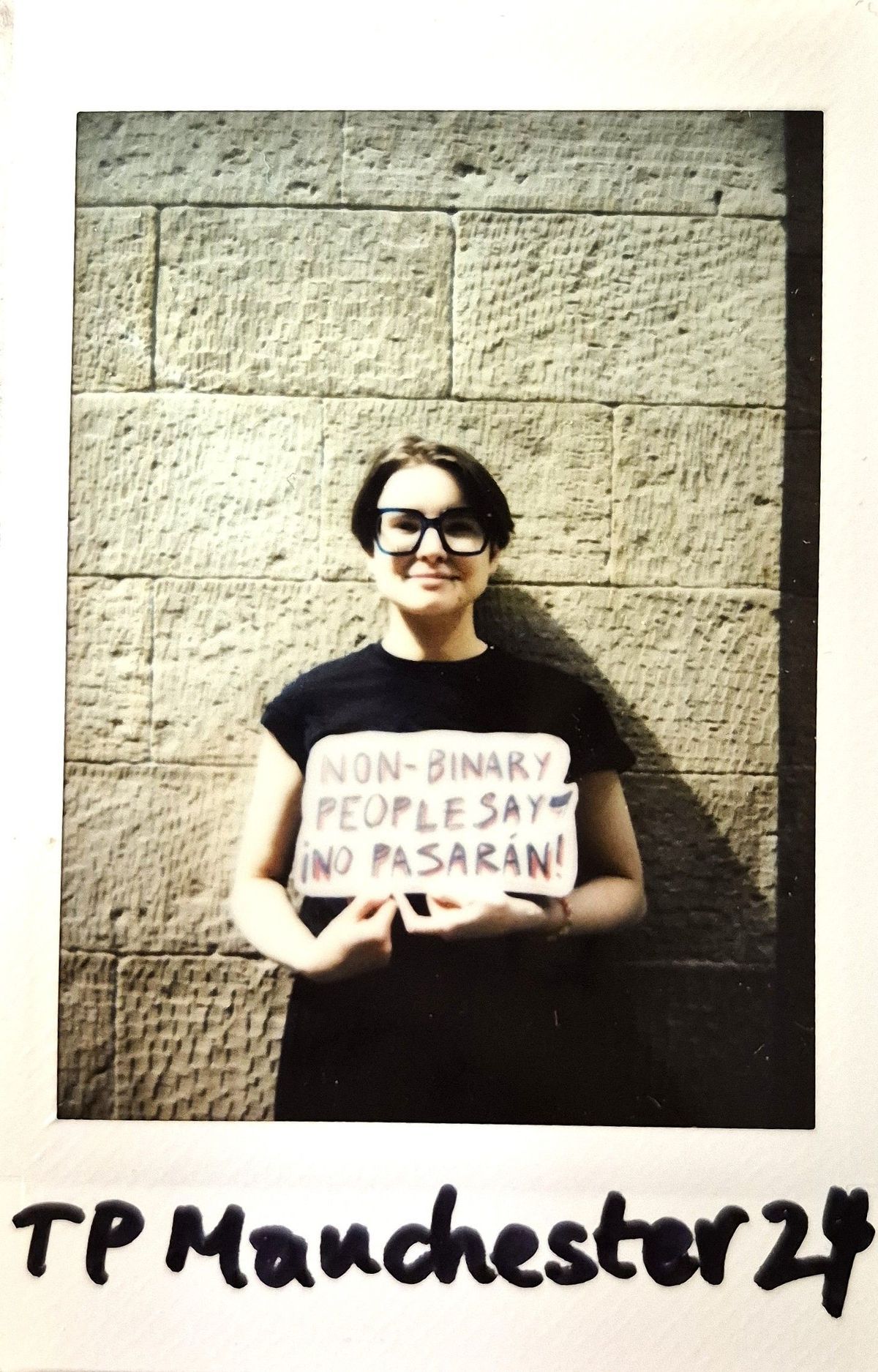 A person holds a sign reading, "Non-binary people say ¡No pasarán!" against a wall.