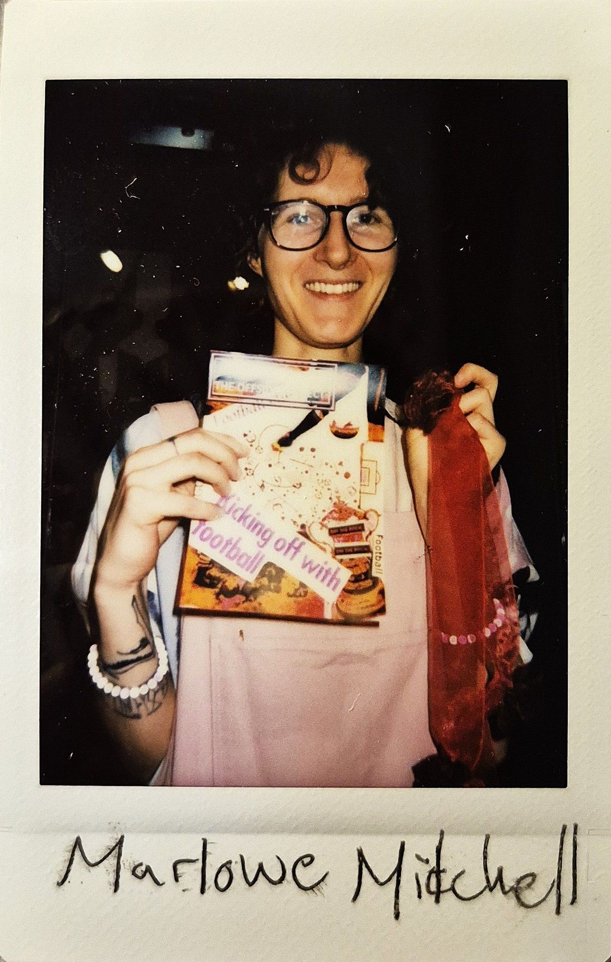 A person is smiling while holding a book and a red fabric ribbon, wearing glasses and bracelets.