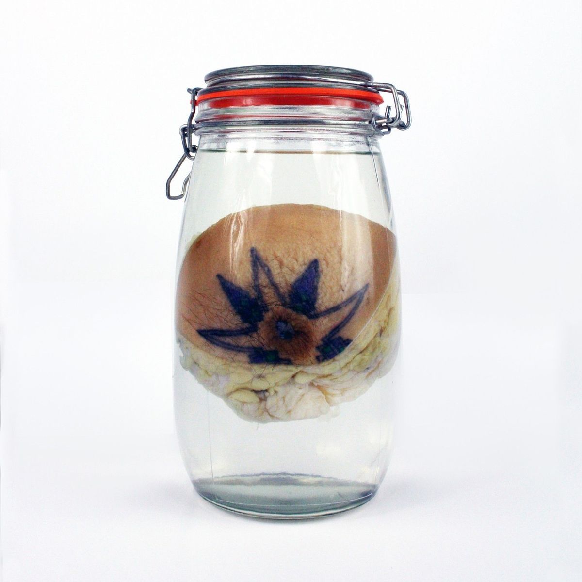 A mason jar which is full of clear liquid with one chest tissue suspended inside. The chest tissue shows a nipple which has a tattoo around it of a star/flower shape.