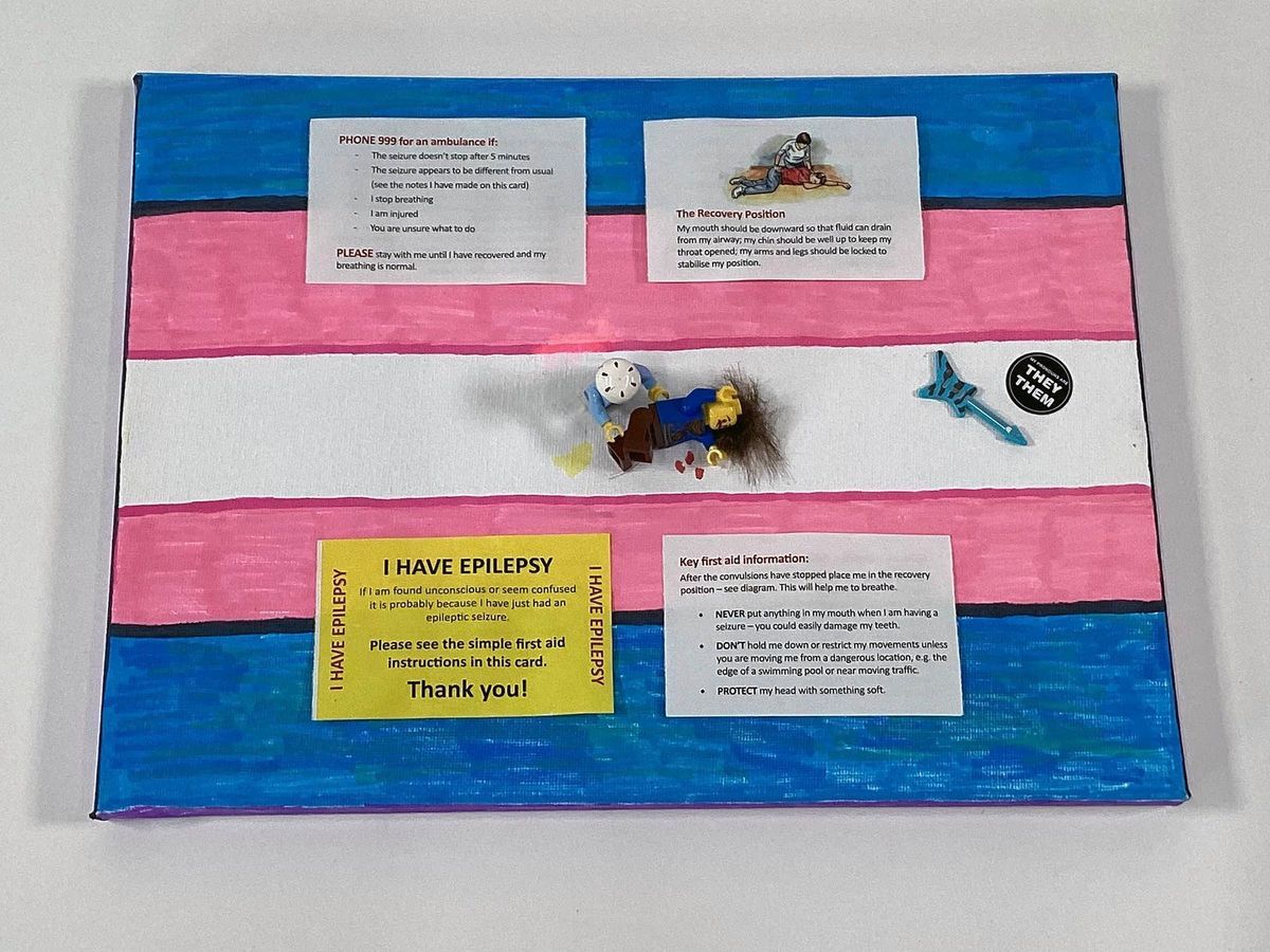 The image shows a canvas, colored in the the trans pride colours,  with epilepsy information, including emergency instructions. There are two Lego figures demonstrating the recovery position.