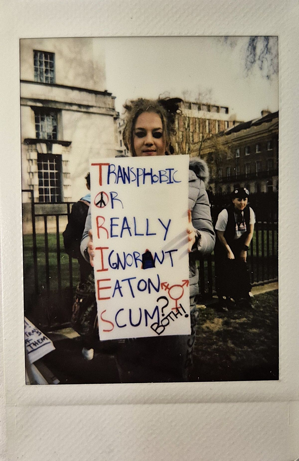 A person holds a protest sign which says "TRANSPHOBIC OR REALLY IGNORANT EATON SCUM? BOTH!"