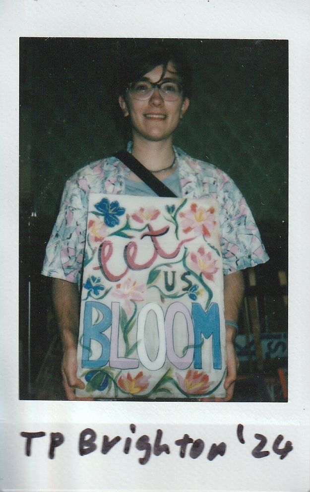 A person holding a colorful floral-themed sign reading "Let Us Bloom," smiling in a casual setting.