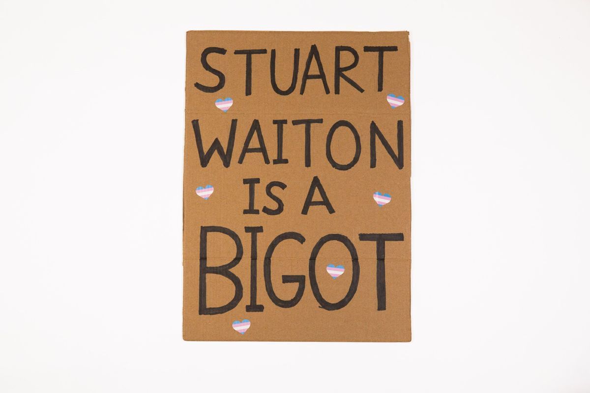 A placard whicch says 'Stuart Waiton is a Bigot' with small trans coloured heart stickers stuck on it.