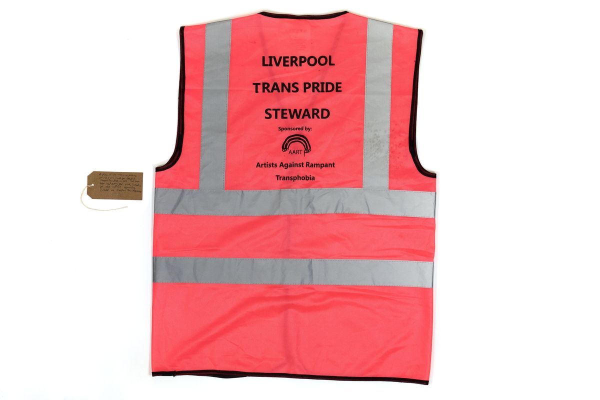 A pink Hi-Vis vest with the text "LIVERPOOL TRANS PRIDE STEWARD Sponsored by Artists Against Rampart Transphobia" printed in black on the back. With a brown, handwritten luggage tag.
