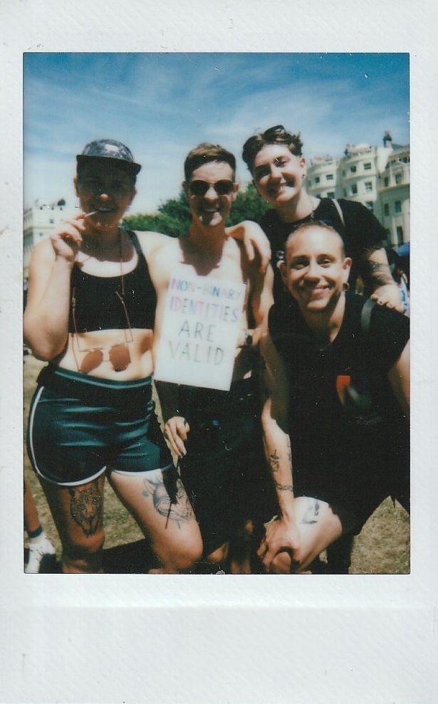 Four people are smiling outdoors, with one holding a sign saying "Non-binary identities are valid."